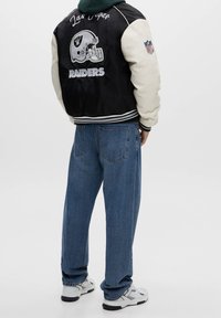 Zwarte en crèmekleurige bomberjack met NFL- en Raiders-logo's, gecombineerd met blauwe spijkerbroek en witte sneakers. De achterkant toont borduurdetails.