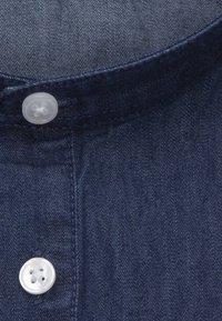 Mörkblå denimskjorta med krage och subtil textur, med en klar plastknapp och kontrasterande sömmar längs kanten.