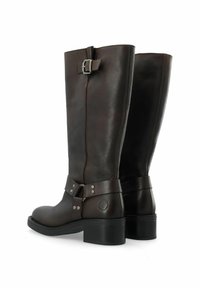 Rudos odinės kelio ilgio botai su plokščiu kulnu, pasižymintys dekoratyvine sagtimi ir dirželio detale su metalinėmis grandinėmis ant kulkšnies.