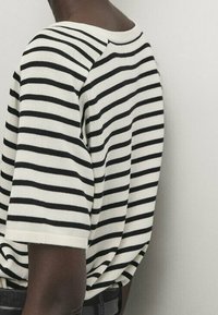 Koszulka krótki rękaw w paski w kolorze kremowym i czarnym, z okrągłym dekoltem i luźnym krojem, z widocznymi szwami i dzianinową fakturą.