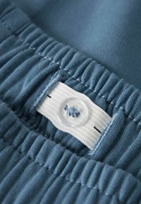 Zils kokvilnas audums ar teksturētu ribu un elastīgu jostu; ir balts tabs ar caurspīdīgu pogu un izšūtu detalizāciju.