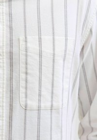 Cămașă albă cu dungi verticale gri, având un buzunar pe piept și un material din bumbac texturat, evidențiind un design casual.