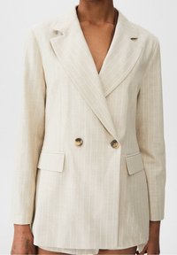 Blazer beige de doble botonadura con rayas blancas, hecho de una tela ligera. Cuenta con solapas con muesca, dos bolsillos frontales y botones redondos.