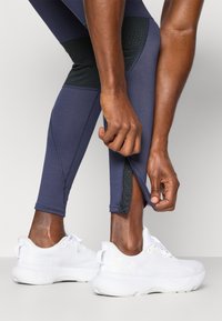 Leggings deportivos de color azul marino con panel texturizado negro, longitud a mitad de pantorrilla y detalle de cremallera en el tobillo; combinados con zapatillas blancas.