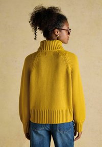 Frau mit lockigem Haar in einem gelben gestrickten Rollkragenpullover und blauen Jeans, seitlich gegen einen beigen Hintergrund posierend.