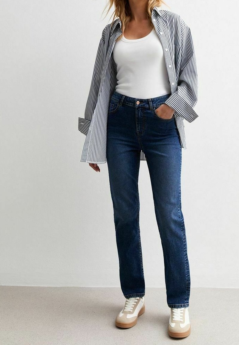 Blå denimjeans med rak passform, kombinerat med en vit linne och en randig skjorta med knappar, avslutat med beige och vita sneakers.