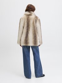 Manteau en fausse fourrure beige et marron avec un col, associé à un jean bleu taille haute à jambes larges et des chaussures noires. Fond minimaliste.