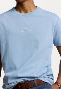 Man wearing light blue Polo Ralph Lauren t-shirt, brown leather belt, denim jeans, and brown leather watch strap, against a plain background.