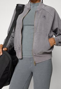 Giacca bomber grigia con zip frontale, polsini a costine e logo ricamato. Indossata sopra un top a costine abbinato e leggings, con una borsa nera.