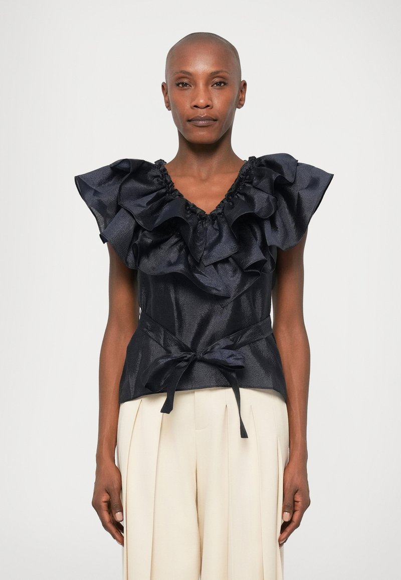 Marineblå silketop med store flæsesleeves og en bundet taljedetalje. Har en V-hals og glat, skinnende overflade. Kombineret med lyse bukser.