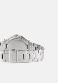 Orologio da polso in metallo argento con bracciale liscio e lucido, cassa rotonda e un design sul retro minimalista con dettagli incisi e branding.