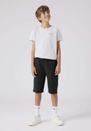 Junge trägt ein hellgraues T-Shirt mit dem Schriftzug "T.W.S.N.", schwarze knielange Shorts, weiße Socken und weiße Turnschuhe, steht vor einem einfarbigen Hintergrund.