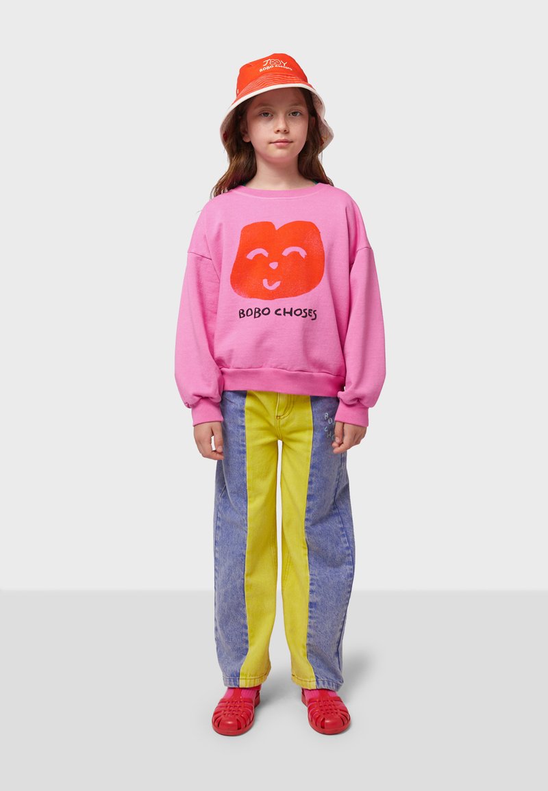 Pink sweatshirt with a red graphic face, paired with blue and yellow split jeans. Red footwear and an orange bucket hat complete the look.