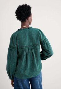 Grün gestreifte Bluse mit Puffärmeln, Rüschenausschnitt und Schmuckleiste auf der Taille. Hergestellt aus strukturiertem Baumwollstoff.