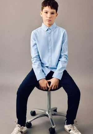 Cămașă albastru deschis cu nasturi, pantaloni bleumarin și teniși albi. Stând pe un scaun rotativ, cu un fundal neted și neutru.