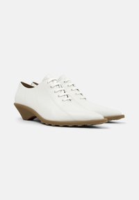 Λευκά δερμάτινα παπούτσια με χαμηλό, γωνιώδες μπεζ τακούνι. Διαθέτουν κορδόνια, μύτη με μυτερό σχήμα και διακριτικές ραφές ως λεπτομέρειες.