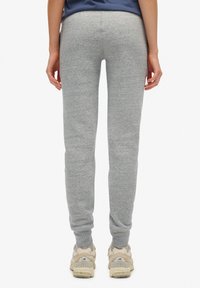 Pantalones de chándal grises hechos de una tela suave, con un corte ajustado y puños elásticos. Llevados con zapatillas de color claro, vistos desde detrás.