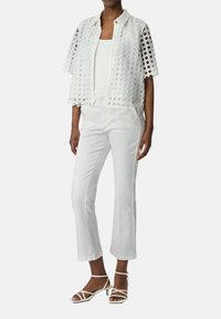 Witte kanten jas met open ontwerp, gedragen over een witte tanktop, in combinatie met witte flared broeken en witte sandalen met bandjes.