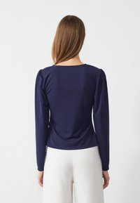 Mørkeblå langermet topp med en tætsittende design, som har rynkede ermer og en rund halslinje, laget av et glatt, elastisk stoff.