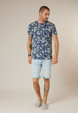 Homme avec des tatouages portant un T-shirt bleu marine à motifs floraux, un short en denim bleu clair retroussé et des chaussures grises sans lacets, debout, regardant vers le bas.