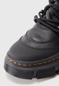 Gros plan d'une botte en cuir noir montrant un dessus texturé, une semelle épaisse avec des coutures jaunes et des lacets noirs sur un fond uni.