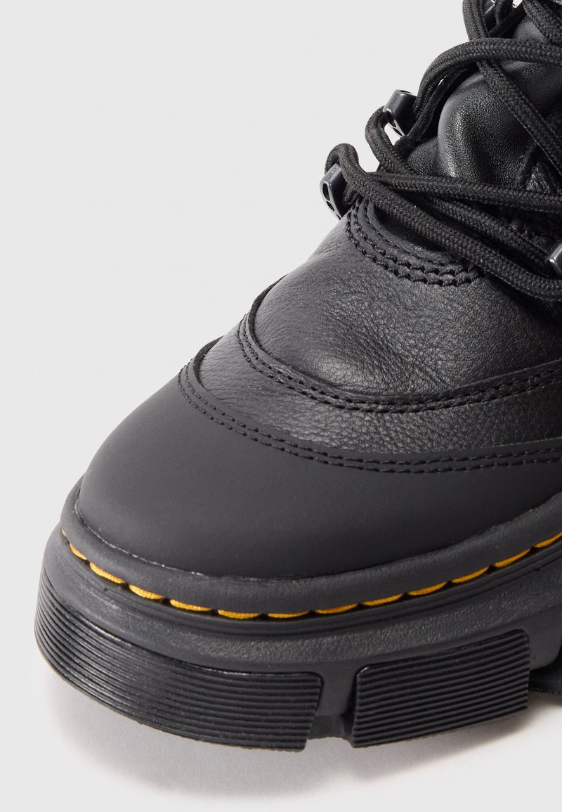 Primo piano di un stivale in pelle nera che mostra la parte superiore testurizzata, la suola spessa con cuciture gialle e i lacci neri su uno sfondo semplice.