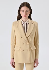 Blazer beige doppiopetto con grandi bottoni rotondi e una texture liscia, abbinato a una camicia di colore chiaro con bordo scuro.