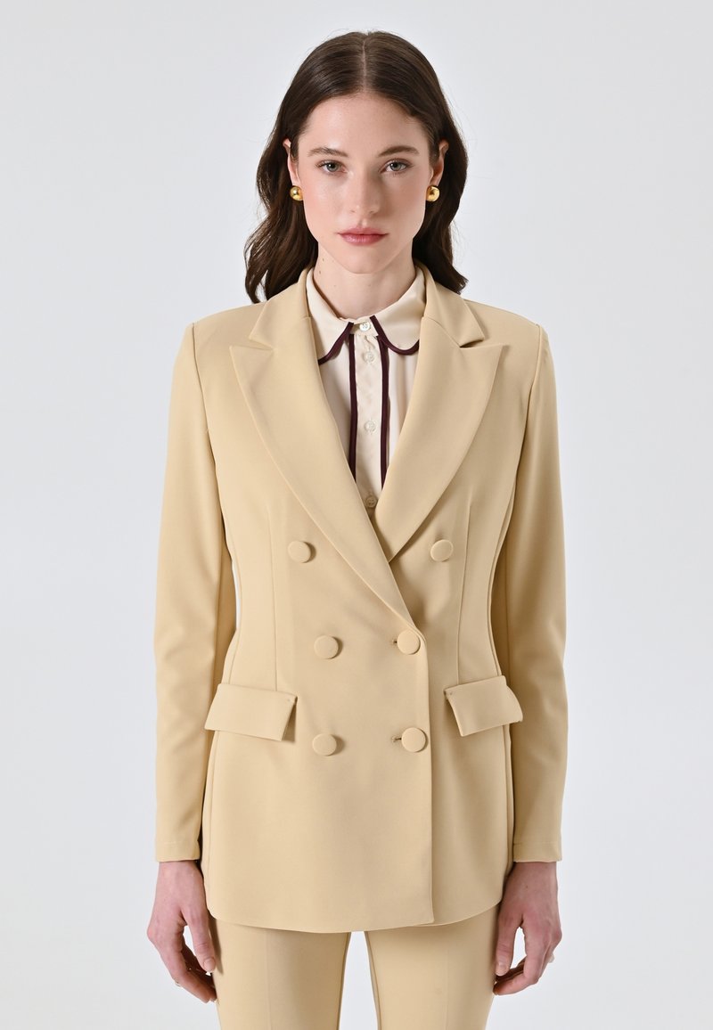 Blazer beige doppiopetto con grandi bottoni rotondi e una texture liscia, abbinato a una camicia di colore chiaro con bordo scuro.
