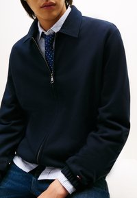 Veste zippée bleu marine à texture lisse et col montant, portée sur une chemise à rayures bleu clair et une cravate à pois, avec des poignets élastiques.