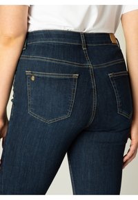 Donkerblauwe jeans met een slim fit, voorzien van achterzakken met zichtbare stiksels en een hoge tailleband. Materiaal lijkt op denim, met een gladde textuur.