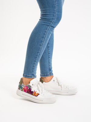 Weiße Turnschuhe mit bunten Blumenmustern, getragen zu blauen Skinny-Jeans, an einer Person, die vor einem weißen Hintergrund steht.