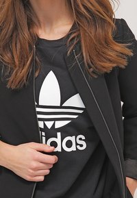 Svart zip-up jacka över en svart T-shirt med en vit Adidas-logotyp och tre ränder mönster. Håret är brunt och vågigt. Hand synlig.