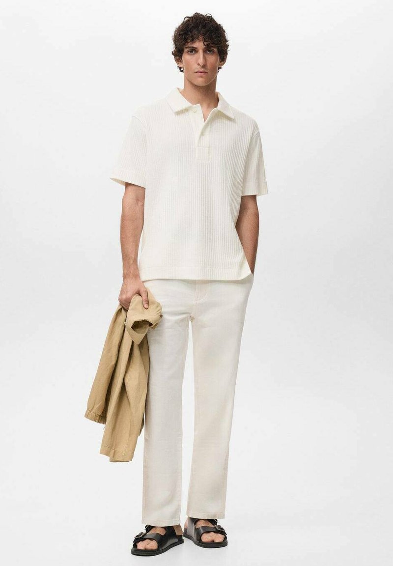 Camisa polo blanca de canalé combinada con pantalones beige claros. El modelo sostiene una chaqueta beige. Las sandalias negras completan el conjunto. Un look sencillo y casual.
