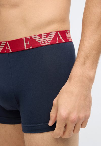 Boxers de algodão azul marinho com uma cintura elástica vermelha com padrão de logotipo. Mão repousando na lateral dos boxers.