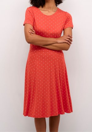 Mujer con los brazos cruzados, vistiendo un vestido rojo de manga corta y largo hasta la rodilla con un patrón de lunares blancos sobre un fondo liso.