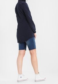 Granatowa koszula z długim rękawem i wysokim kołnierzem, zestawiona z dżinsowymi szortami i białymi trampkami za kostkę. Gładki materiał o dopasowanym kroju.