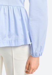 Šviesiai mėlyna palaidinė su peplum juosmeniu, susirinkusiomis ilgomis rankovėmis ir lygiu paviršiumi. Apačia turi tiesią iškirptę. Dėvima ant baltų kelnių.