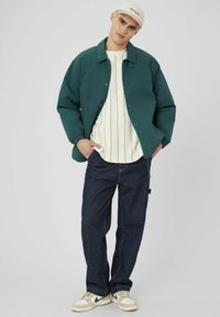 Veste en padded bleu canard, chemise à rayures crème, jeans en denim foncé et baskets beiges avec des accents noirs. Le mannequin pose de manière décontractée avec les mains dans les poches.