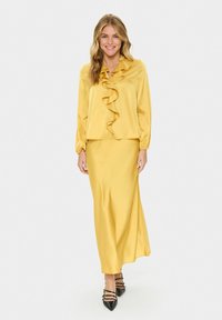 Gelbe Satinbluse mit Rüschen, kombiniert mit einem passenden satin Maxi-Rock. Schwarze Absatzschuhe mit Knöchelriemen. Glatte Textur, elegantes Design.