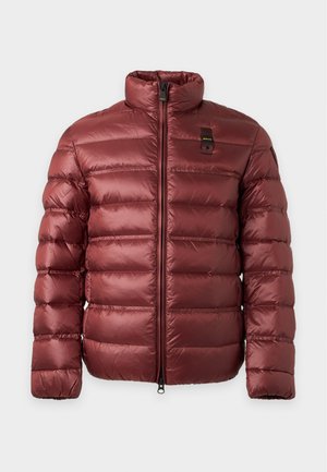 Jeune homme debout portant une veste matelassée rouge par-dessus un t-shirt blanc, pantalon noir et chaussures noires sur un fond clair uni.