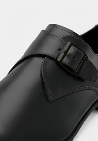 Μαύρο συνθετικό παπούτσι με λεία επιφάνεια, μυτερή μύτη, φαρδιά λωρίδα με ορθογώνια αγκράφα και αντίθετη λεπτομέρεια ραφής.