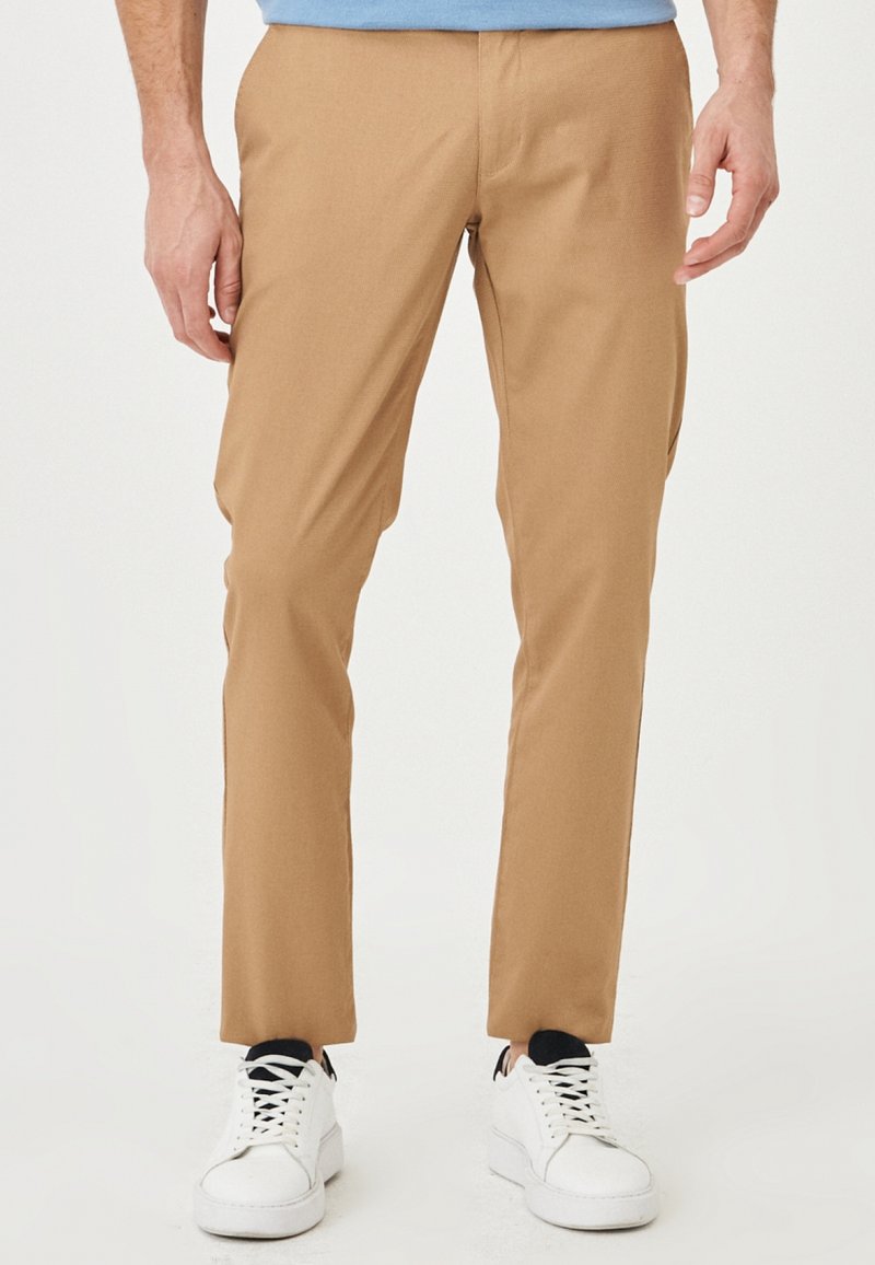 Homme portant un pantalon camel slim, des chaussettes noires, des baskets blanches et une chemise bleu clair partiellement visible, debout devant un fond blanc.