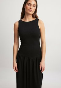 Svart ärmlös klänning med figurnära bodice och draperad kjoldetalj. Tillverkad av texturerad tyg, med rund halsringning och subtil lyster.