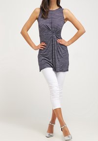 Túnica gris sin mangas con detalle retorcido en la cintura, combinada con leggins blancos recortados y zapatos de tacón plateados sobre un fondo neutro.