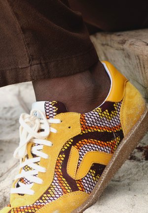 Pied portant une chaussure de sport jaune et marron à motifs avec des lacets blancs, reposant sur une surface sablonneuse à côté de marches en bois usées.