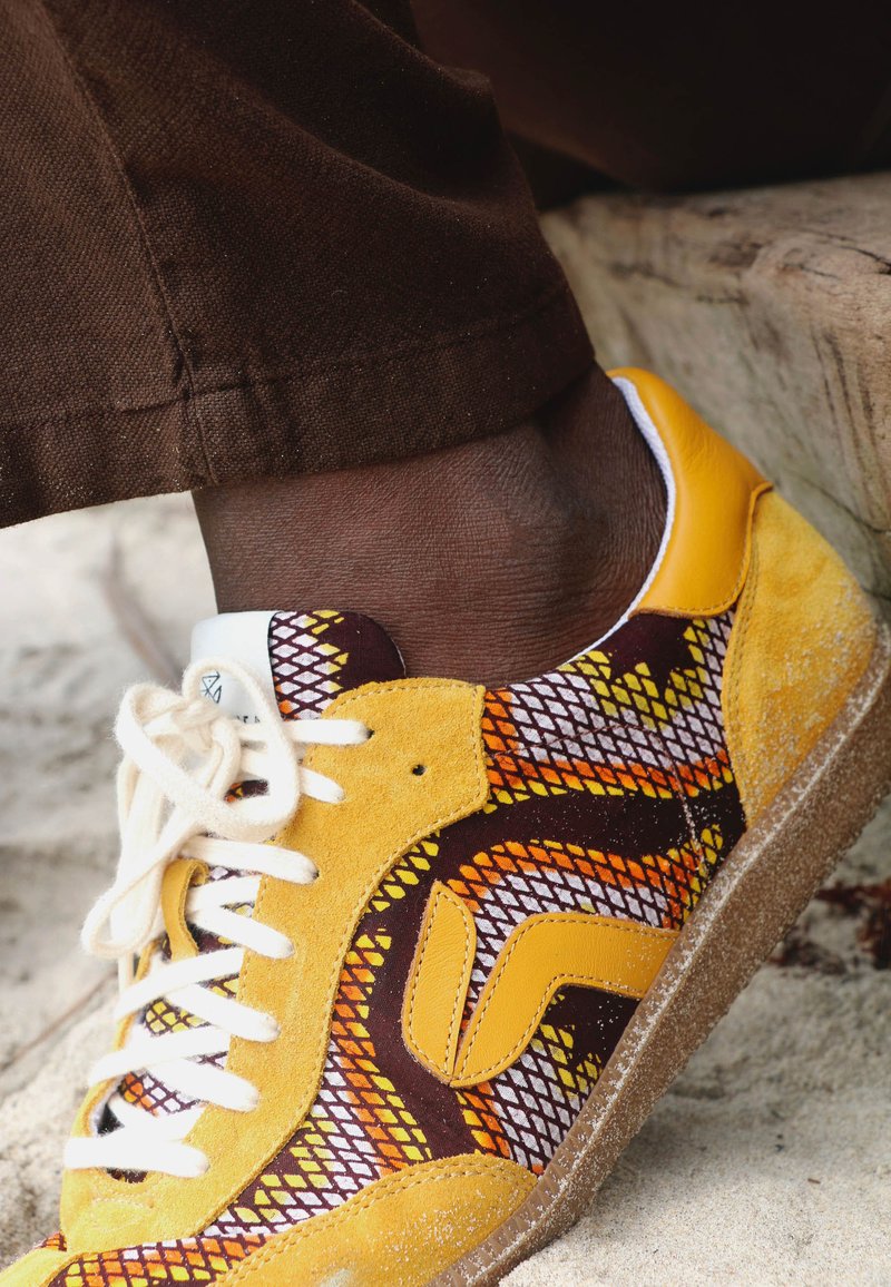 Pied portant une chaussure de sport jaune et marron à motifs avec des lacets blancs, reposant sur une surface sablonneuse à côté de marches en bois usées.