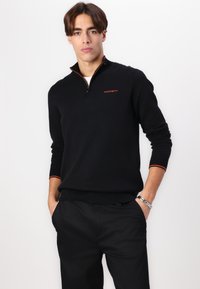 Sudadera negra con cremallera y acentos naranjas, con un logo en el pecho. Mangas y dobladillo acanalados. Textura suave, diseño ajustado, llevada sobre pantalones oscuros.