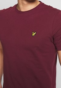 Bordová bavlněná tričko s kulatým výstřihem. Na levém prsu se nachází malé žluté vyšívané logo ptáčka. Hladká textura.