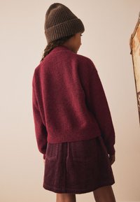 Pulover tricotat bordo cu croială lejeră, asortat cu o fustă din catifea côtelée în aceeași nuanță, prevăzută cu buzunare frontale și textură striată.