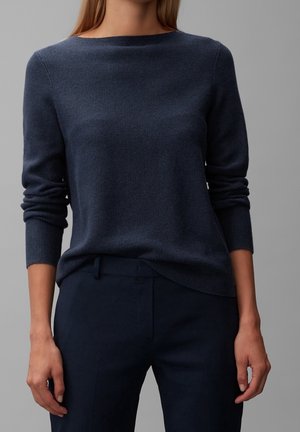 Femme portant un pull à manches longues texturé bleu marine et un pantalon bleu marine assorti sur un fond gris uni.